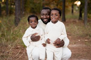 A smiling man poses outdoors with two young boys in matching cream outfits on a wooded path.