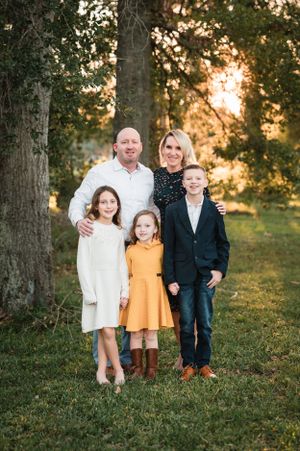 Outdoor family portrait with coordinated outfits standing together among trees at golden hour.