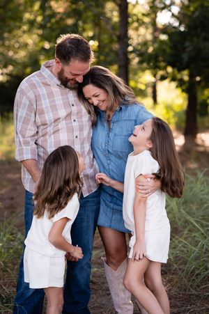Family of four smiling and laughing together outdoors in a wooded setting, parents and two young girls in coordinating outfits.