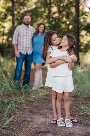 Two young girls hugging outdoors with parents standing behind them in a wooded forest family portrait.