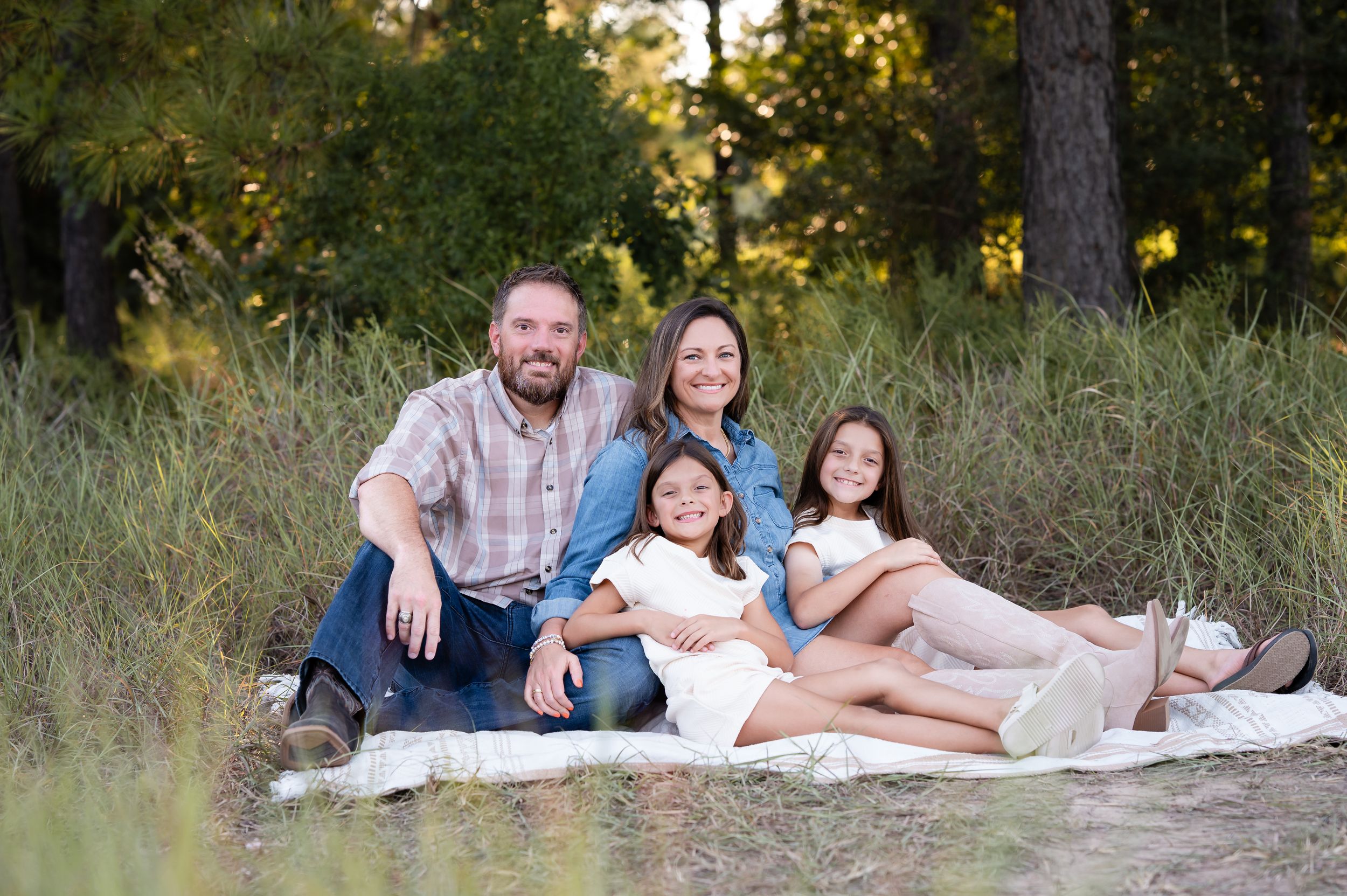 Family of four smiling together on a blanket in a lush green field surrounded by tall pine trees.