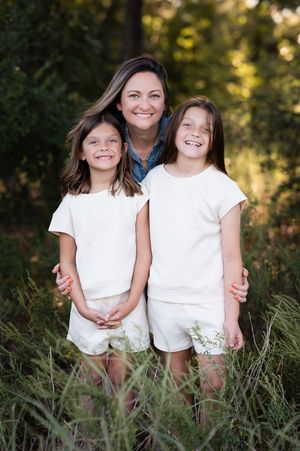 A smiling woman poses outdoors with two young girls in matching white outfits, surrounded by lush green foliage.