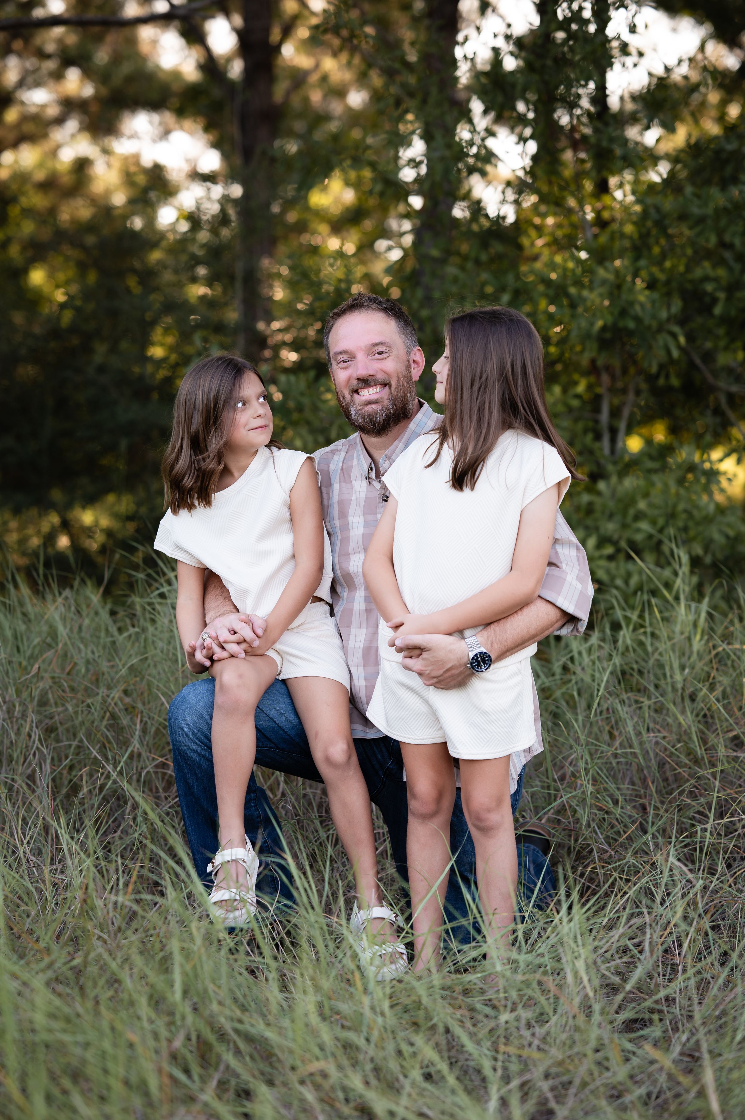 A smiling man sits outdoors in tall grass with two young girls in white dresses beside him, surrounded by green trees.
