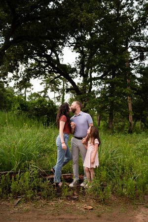 Family of three sharing a tender moment outdoors, standing on rustic railroad ties surrounded by lush green trees.