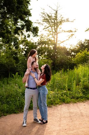 Joyful family photo at golden hour, dad carrying daughter on shoulders while mom smiles in a sunlit park.