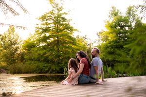 Family of three sitting on a wooden dock by a serene pond surrounded by lush green trees in golden sunlight.