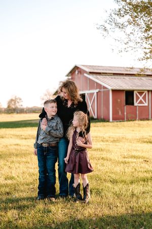 A mother and two children pose in a golden field near a red barn during a warm family portrait session.