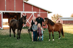 Family of four poses with two horses in front of a red barn on a sunny day at a rural farm.