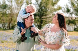 Happy family with toddler on dad's shoulders smiling outdoors in a blooming wildflower field.