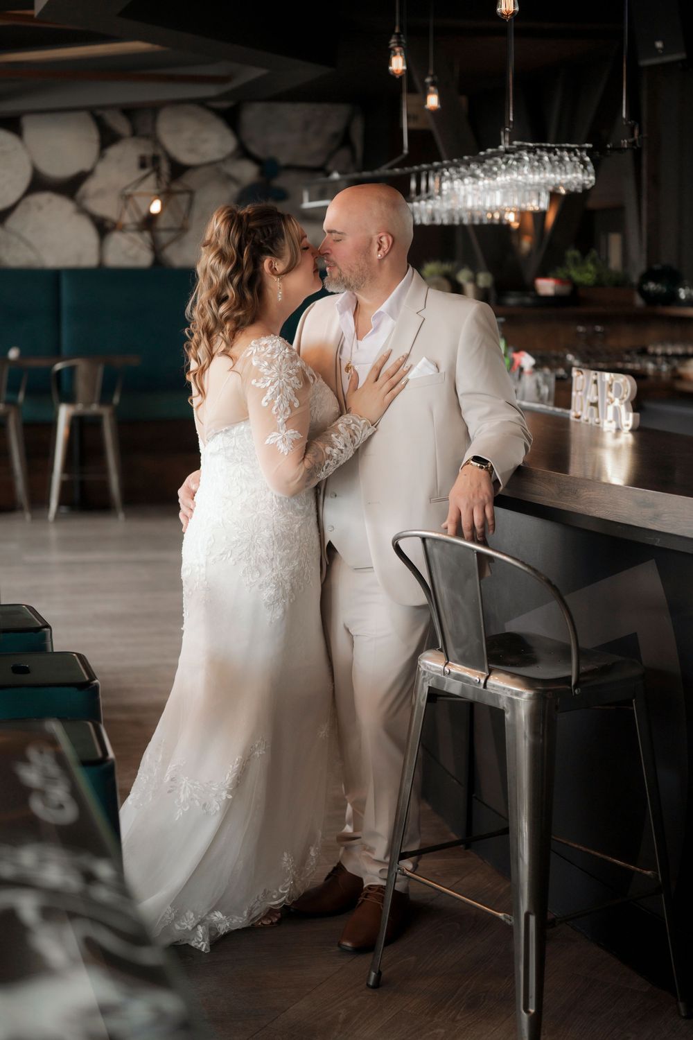 A couple in wedding attire share an intimate moment at a modern bar counter with hanging pendant lights.