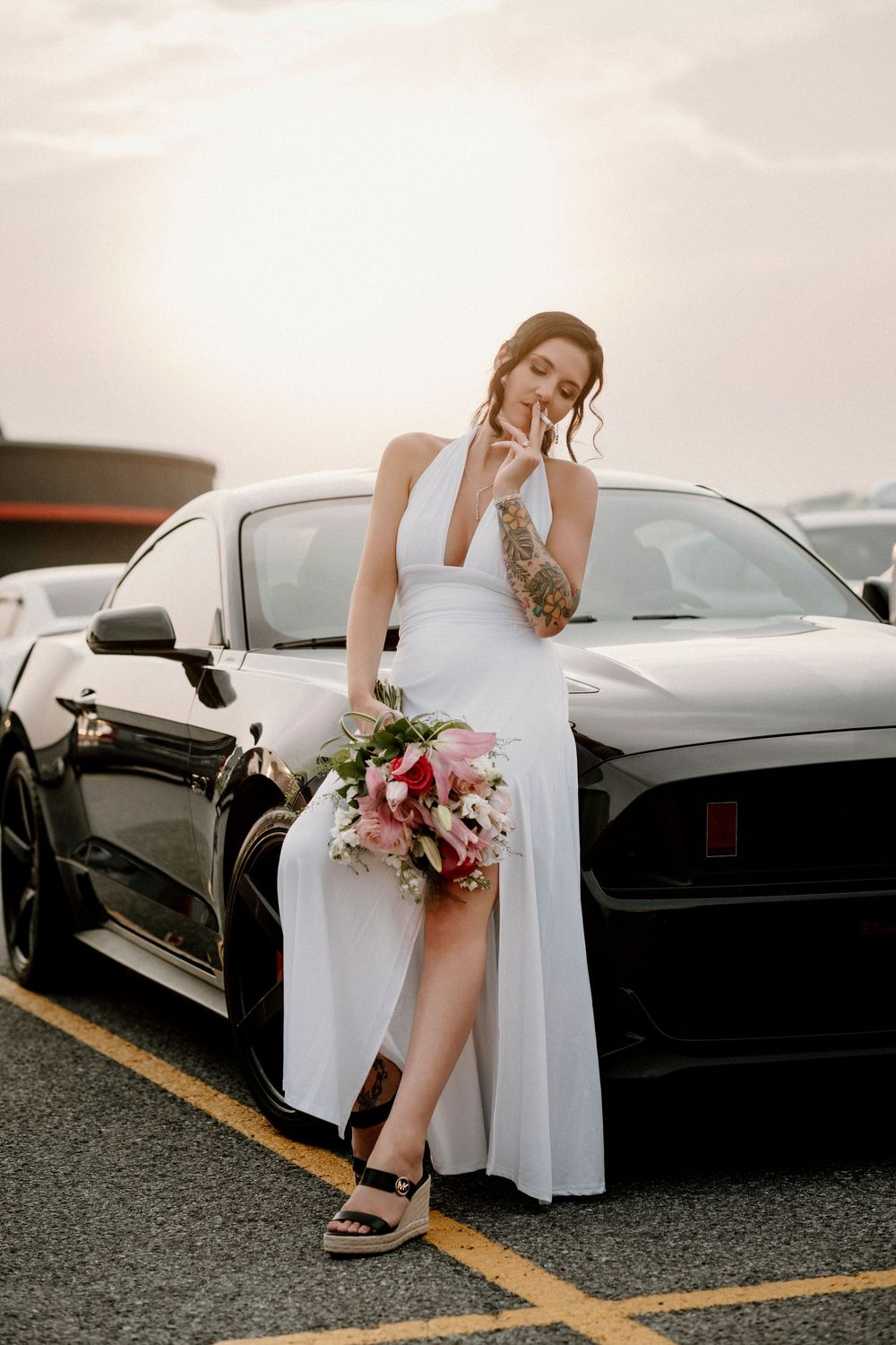 A figure in a white dress sits elegantly against a black luxury car in a parking lot with flowers in hand.