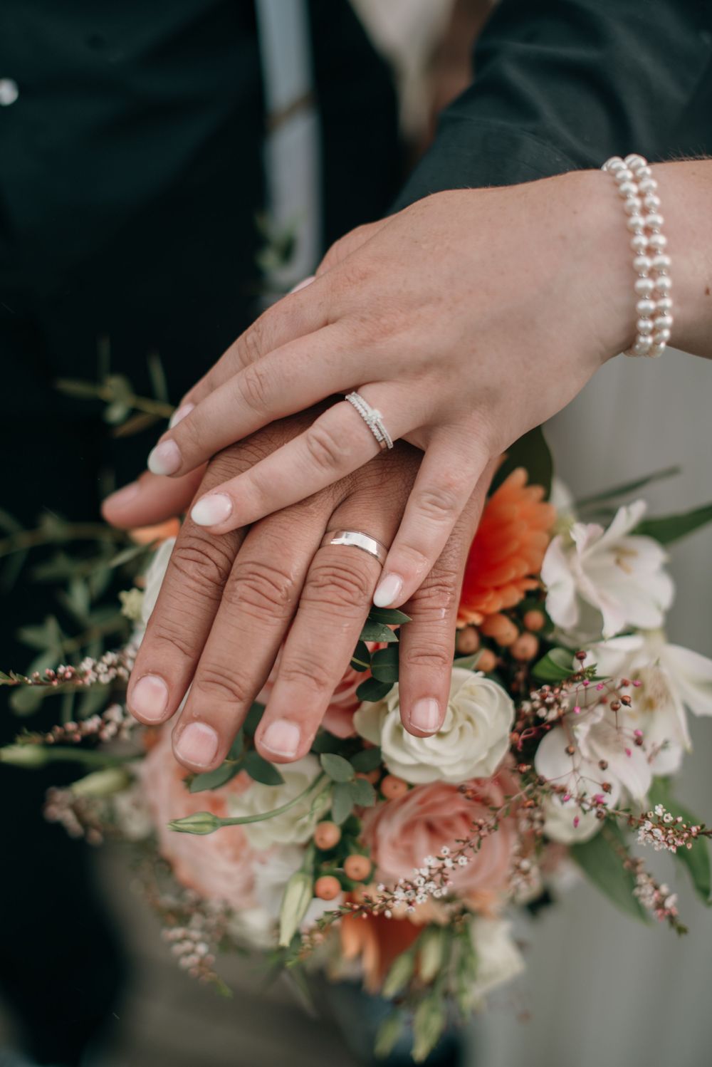 Close-up view of hands wearing wedding rings against a soft floral bouquet with roses and greenery.