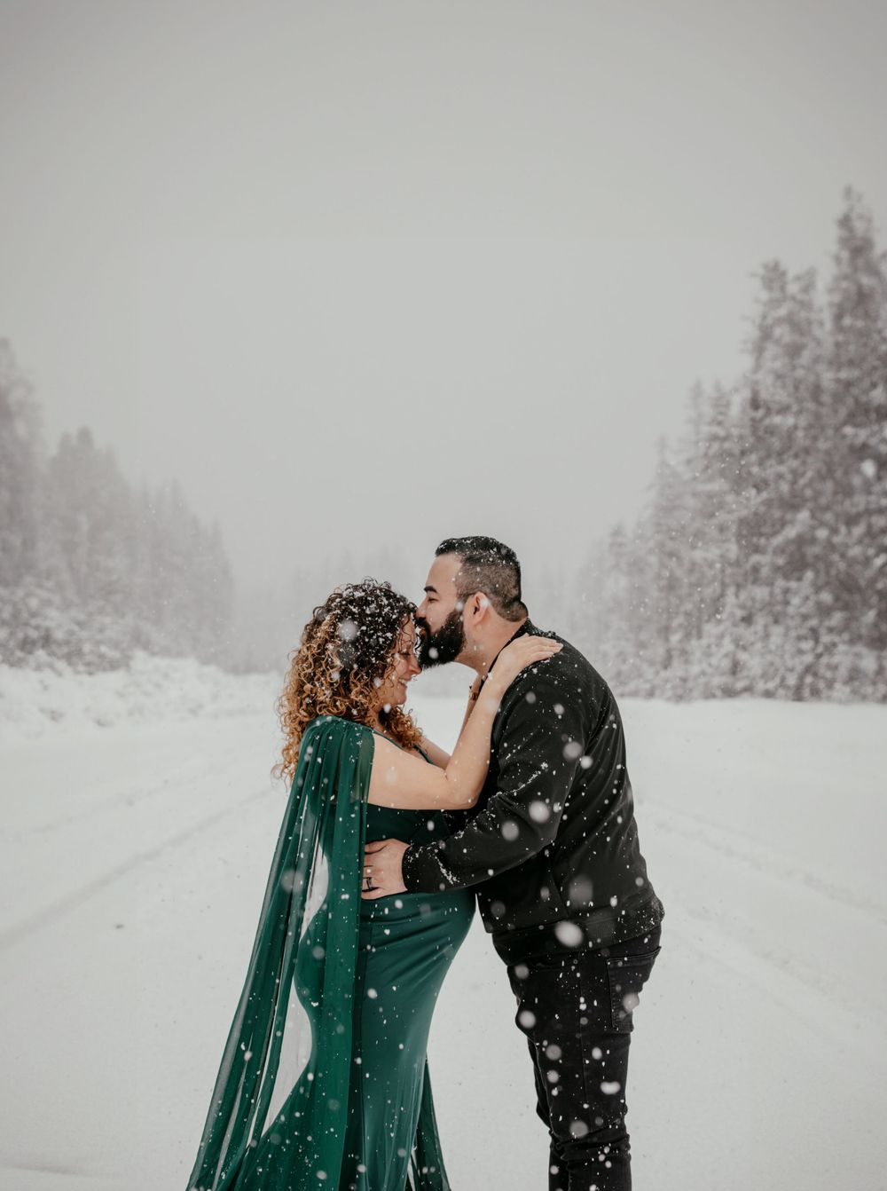 Romantic couple embracing in snowy forest landscape wearing emerald green attire.