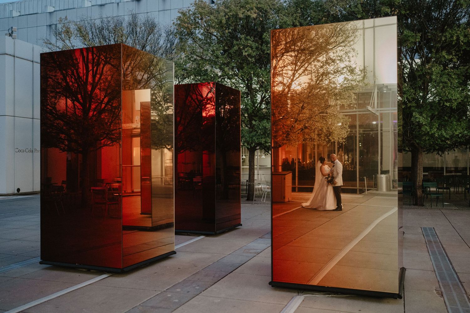 Modern art installation with mirrored panels reflecting trees and sunlight in an urban space creates a warm orange glow.