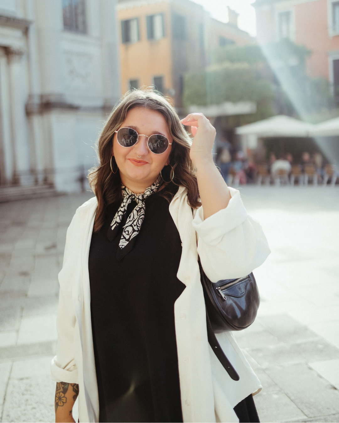 Fashionable woman in black dress and white blazer poses on city street with sunglasses and statement necklace.