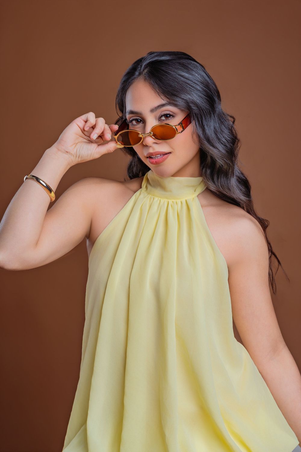 Woman in yellow halter dress lowering amber sunglasses, posing against brown background.