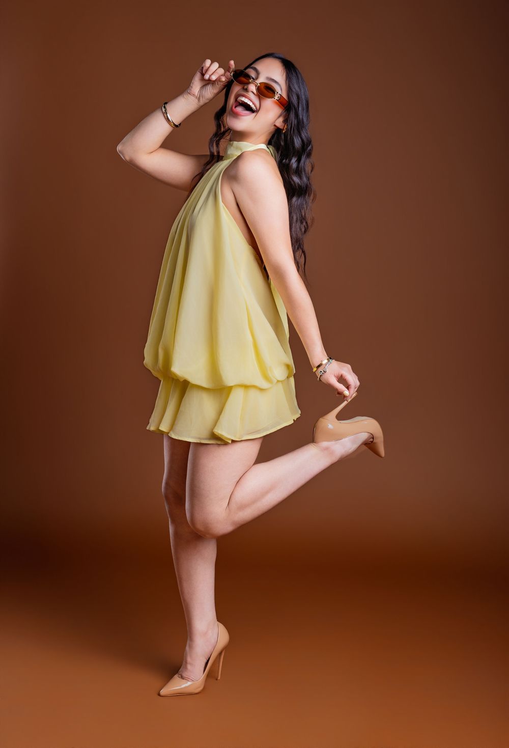 Woman in yellow halter dress and nude heels posing playfully with sunglasses on brown studio background.