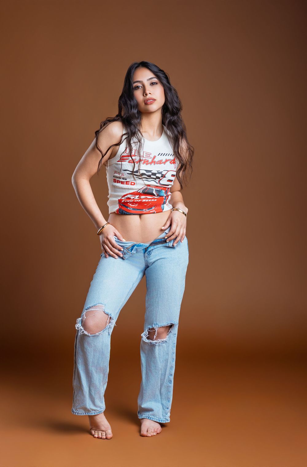 Woman posing in ripped jeans and a NASCAR crop top against a brown studio background.