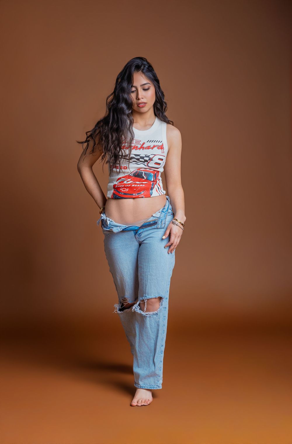 Young woman in ripped jeans and a racing graphic crop top poses barefoot against a warm brown studio background.