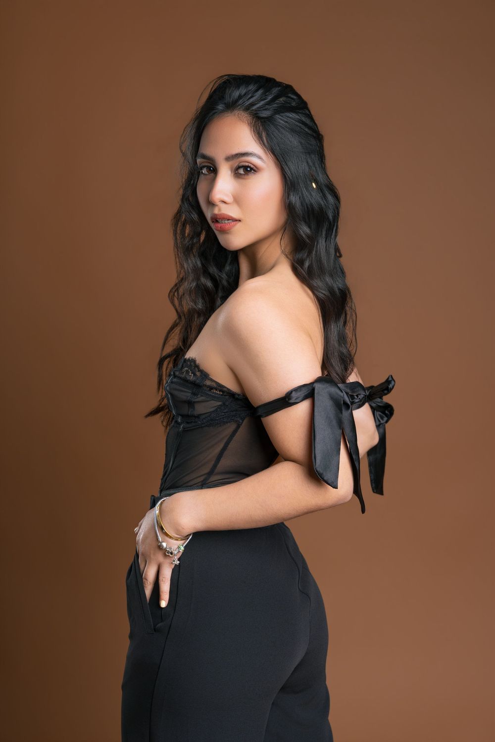 Woman in black off-shoulder corset top with bow details and black pants poses against brown background.