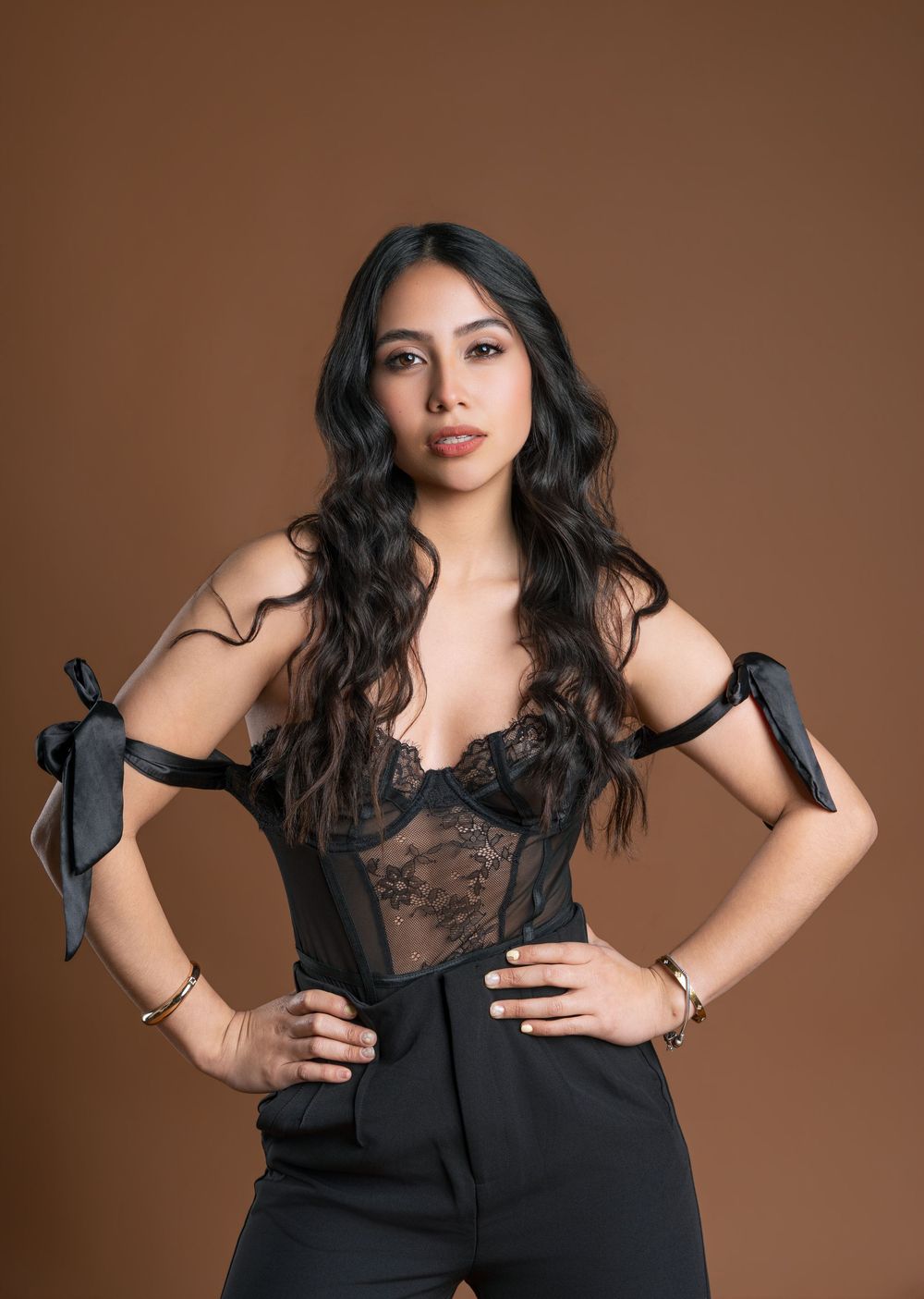 Woman in black lace bodysuit with bow-tie shoulders and black pants poses confidently against a brown background.