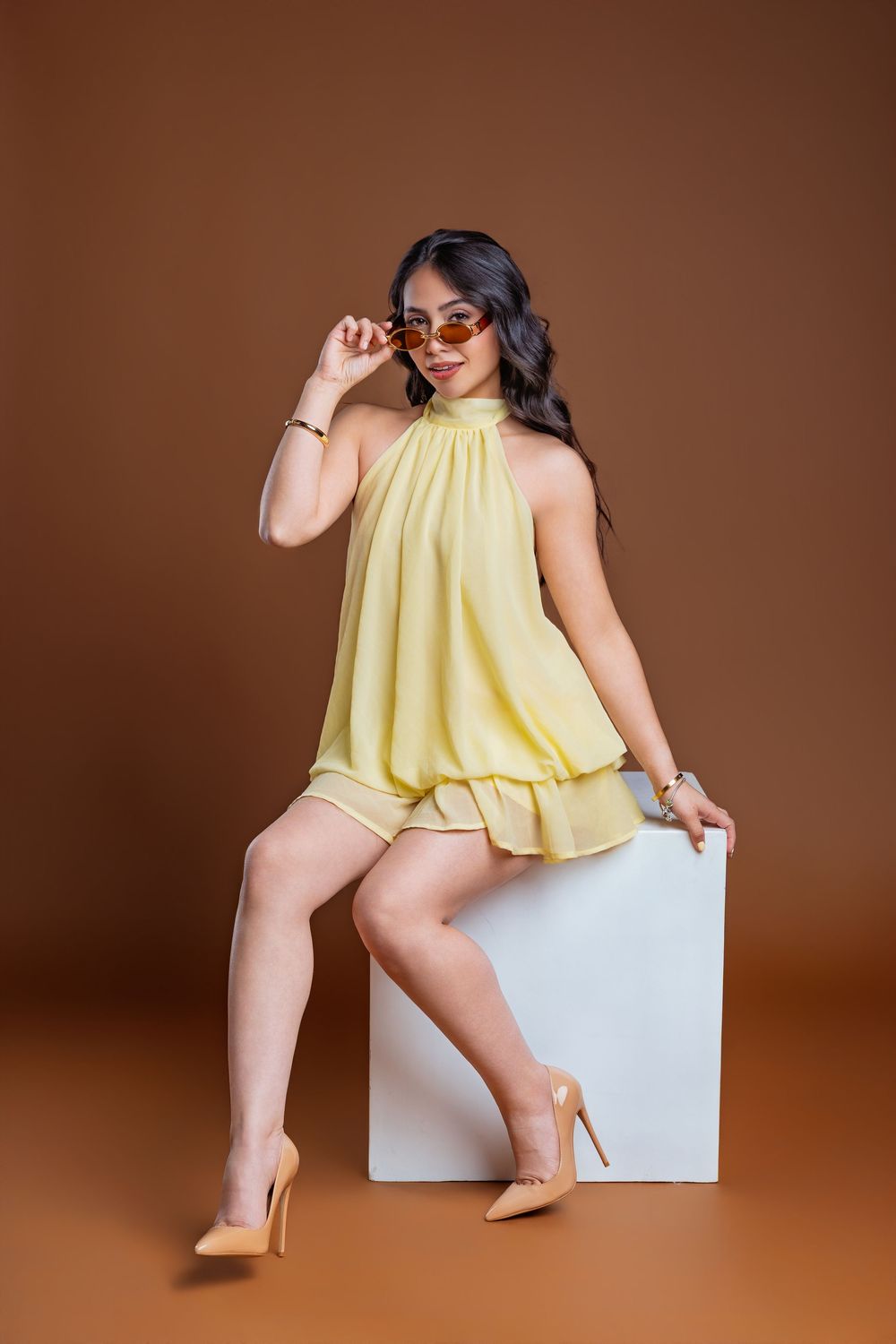 Woman in yellow halter top and shorts, sitting on white cube, wearing sunglasses and nude heels, brown background.
