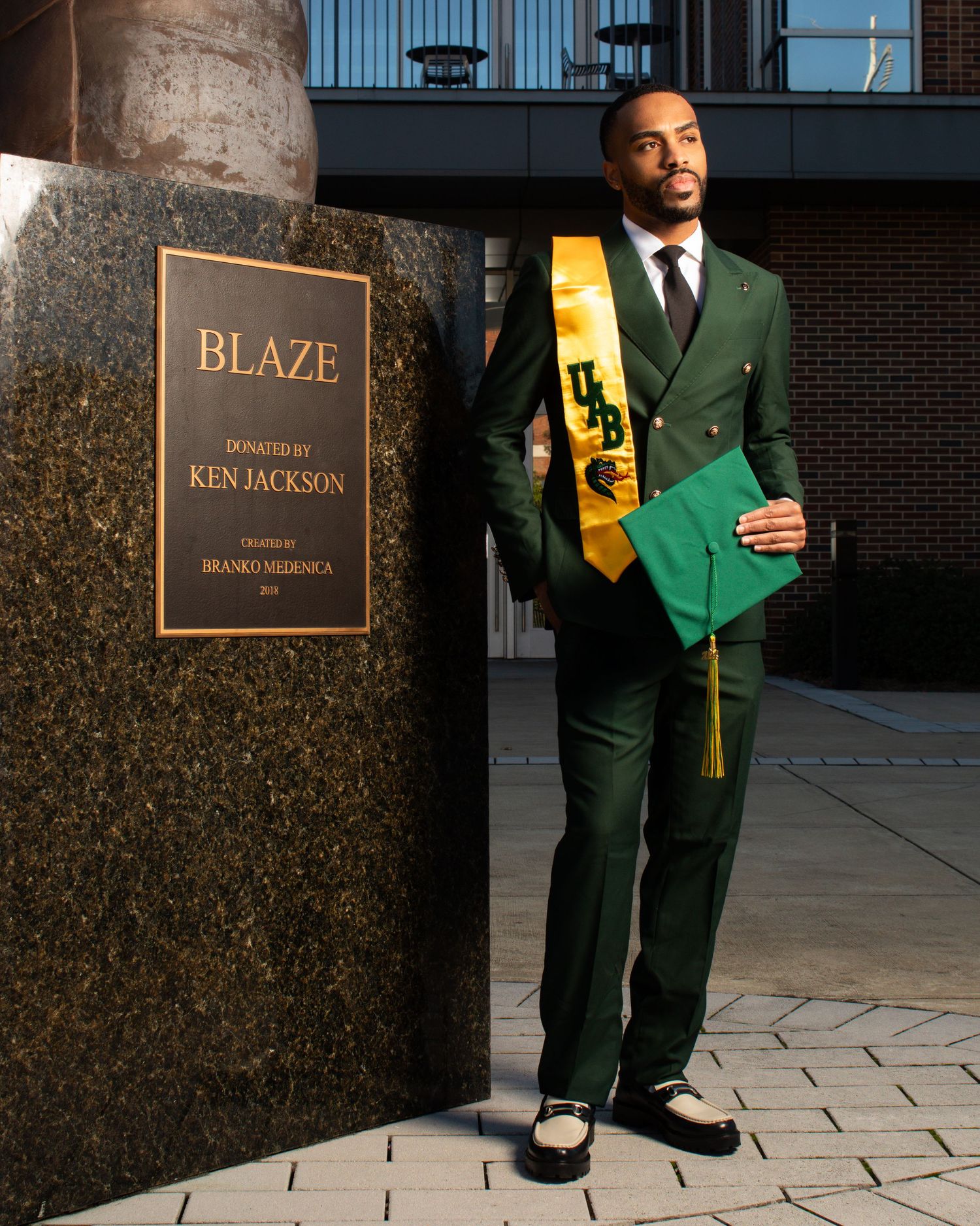 Graduate in green suit with gold sash standing next to Blaze monument sign.