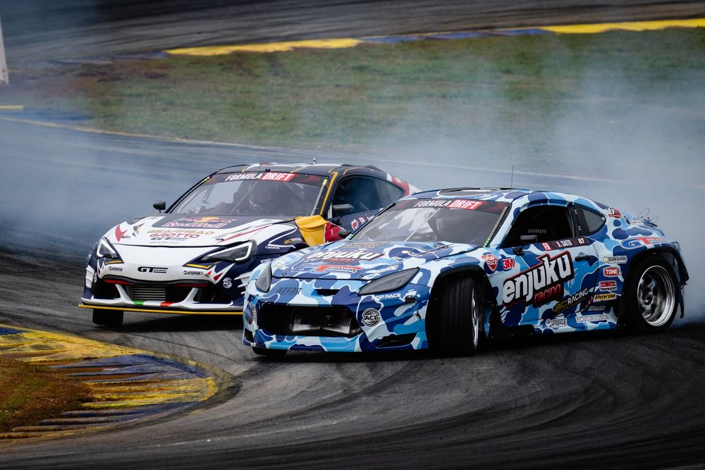 Multiple drift cars sliding in tandem through a race track corner.
