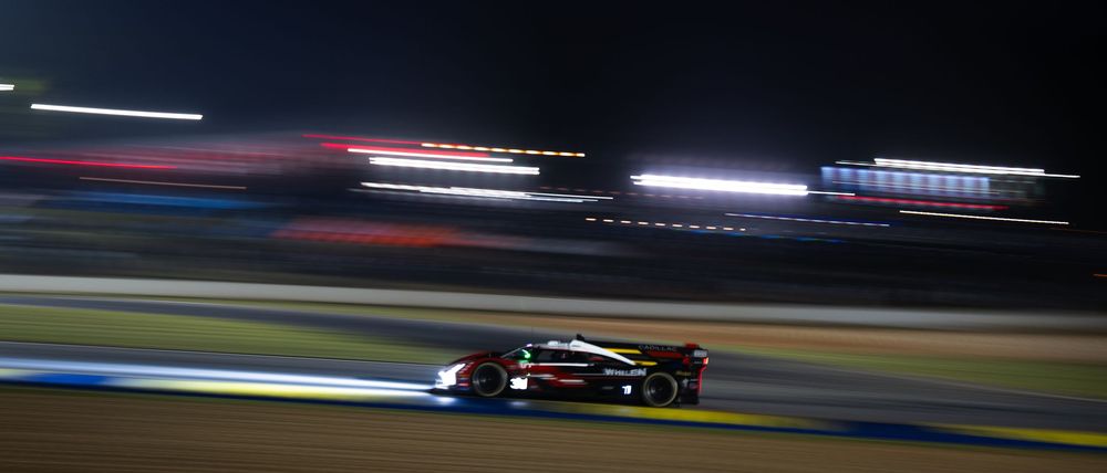 Racing cars speed around a track at night with motion blur and light trails.