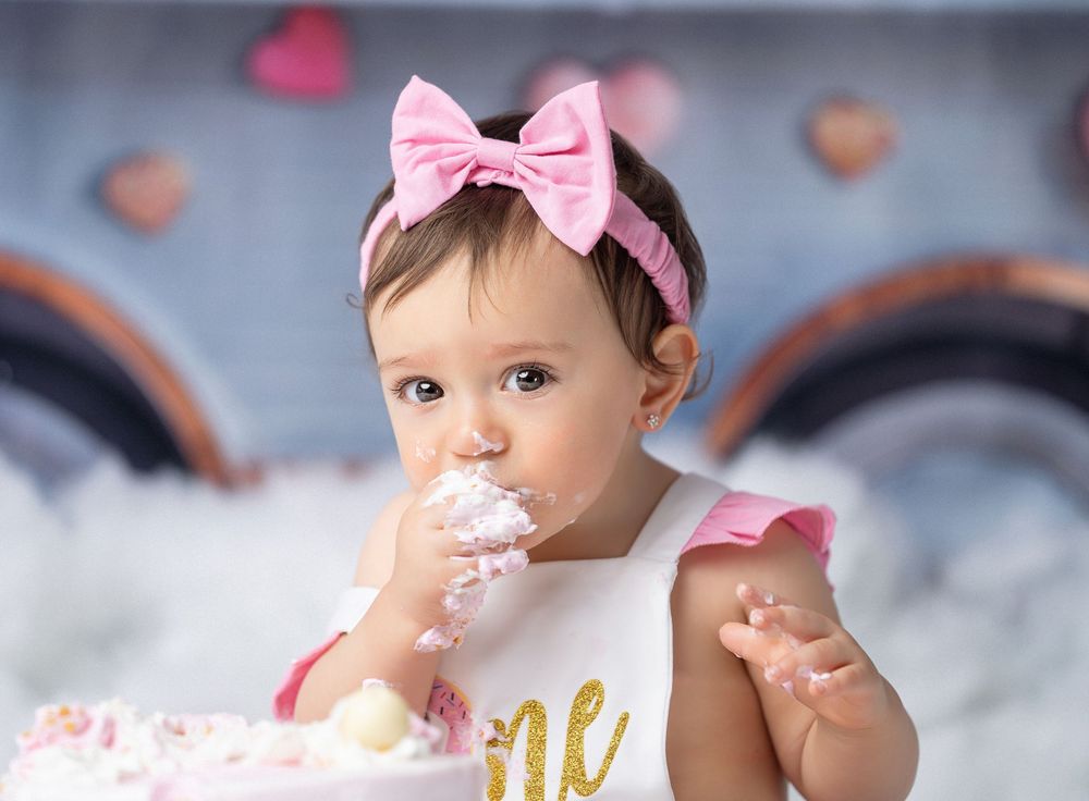 Miami Maternity photographer A baby wearing a pink bow headband enjoys tasting cake at a birthday celebration with a gray background.