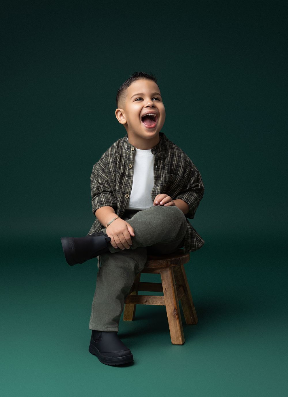 Miami Maternity photographer A joyful young child wearing a plaid shirt and gray pants laughs while sitting on a wooden stool