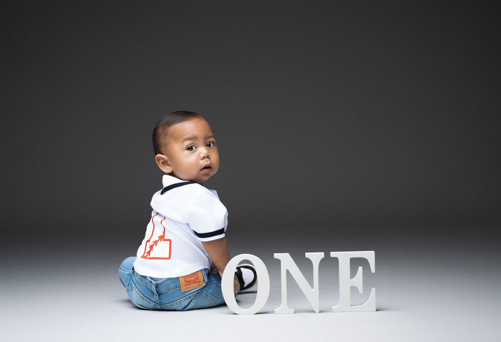 Miami Maternity photoBaby in a white t-shirt sits next to decorative 'ONE' letters during a milestone photo session against dark background.