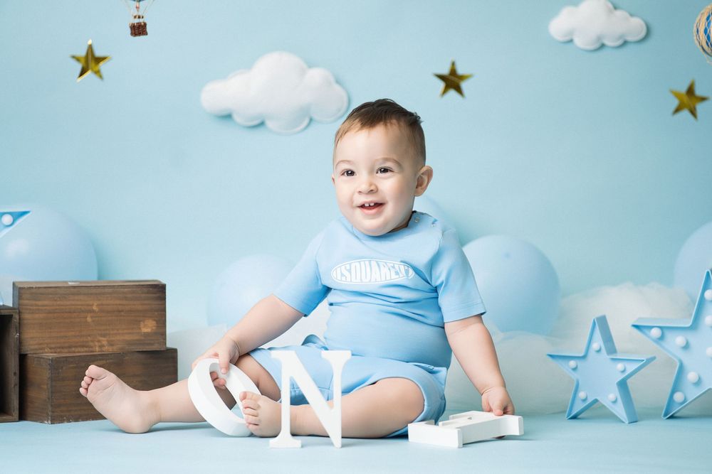 Miami Maternity photographeA baby in light blue outfit sits on blue backdrop with decorative white clouds, wooden box, and star decorations.