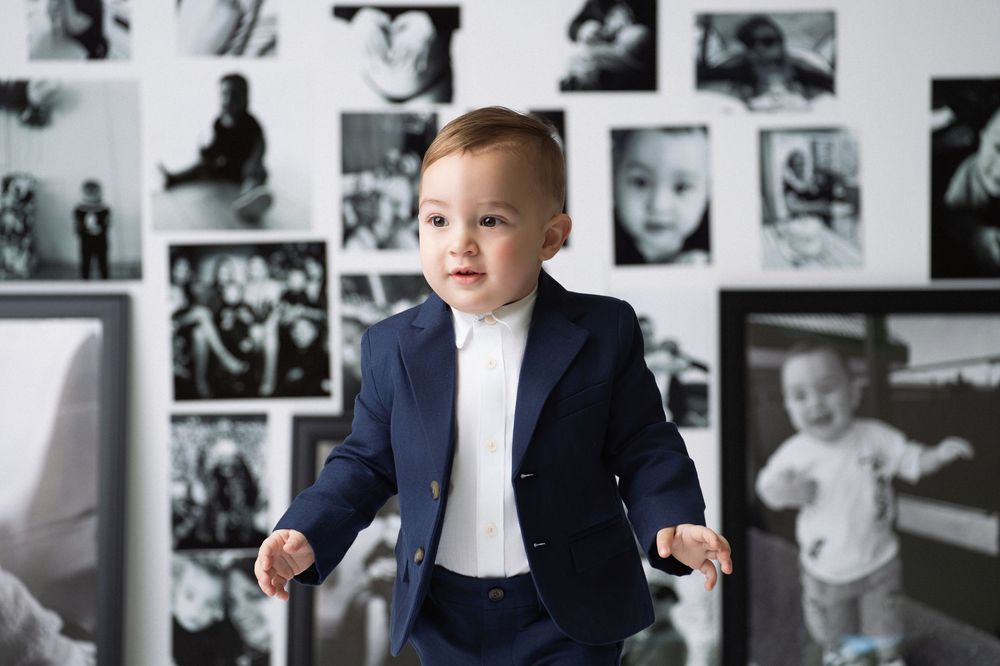 Miami Maternity photogrA young child in a navy blazer and white shirt stands with arms spread out against a wall decorated with photographs.