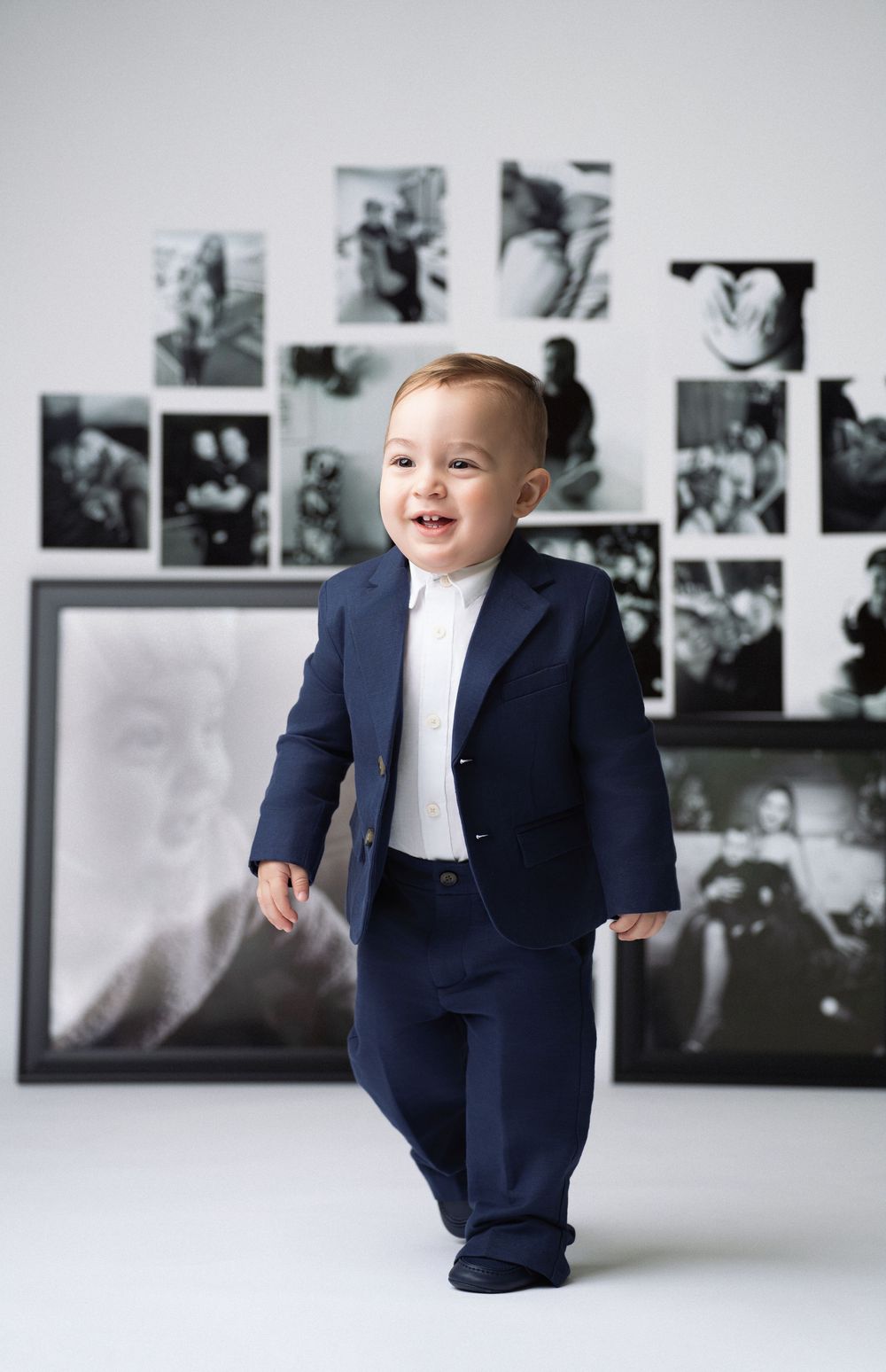 Miami Maternity photographer Baby wearing navy blue suit stands in front of black and white photo gallery wall with bright smile.