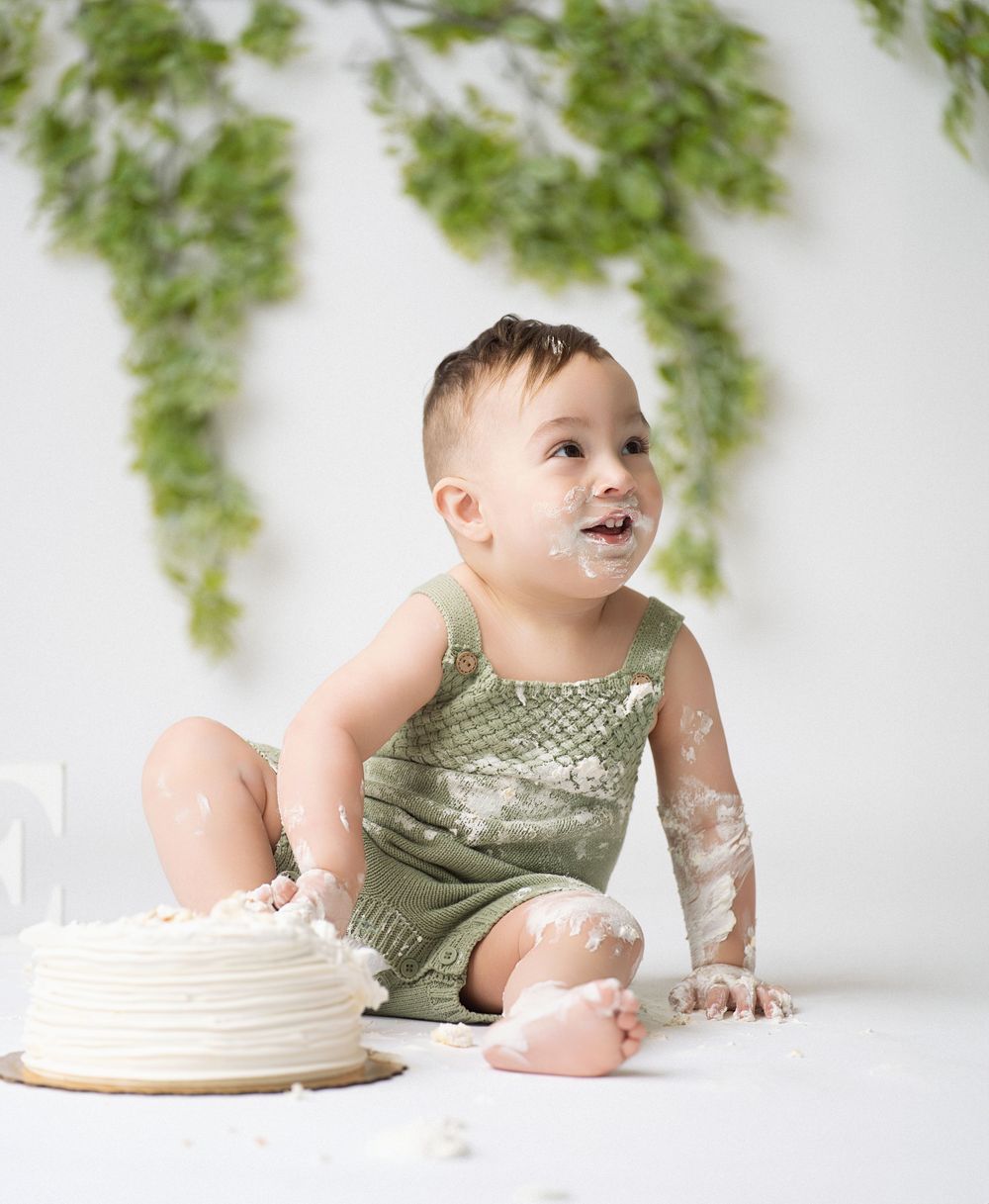 Miami Maternity photographer Sweet one-year-old in green outfit sits with cake and ivy backdrop during messy cake smash photo session.