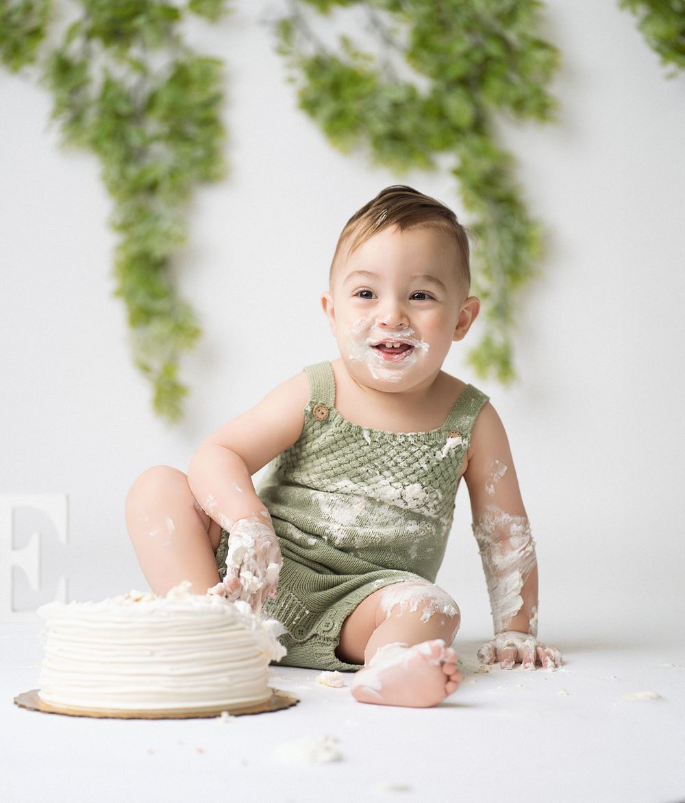 Miami Maternity photographer Baby wearing green romper enjoys smashing and playing with white frosted cake during smash cake photo session.
