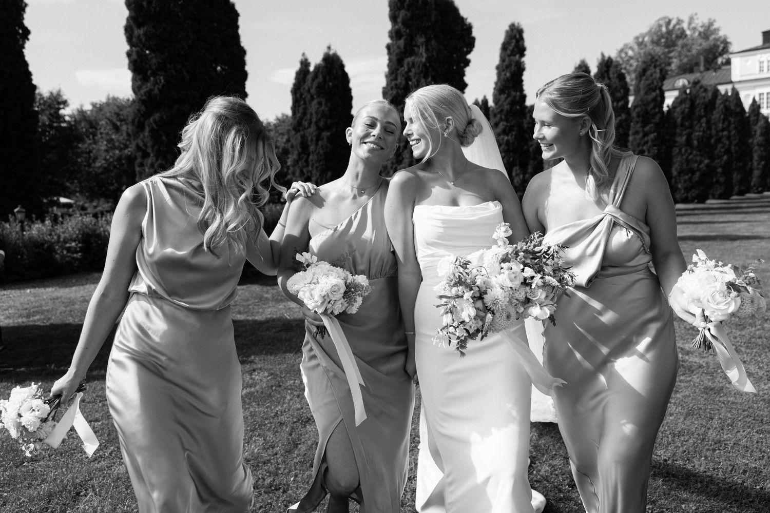 Black and white candid photo of bridesmaids in flowing dresses sharing a joyful moment outdoors.