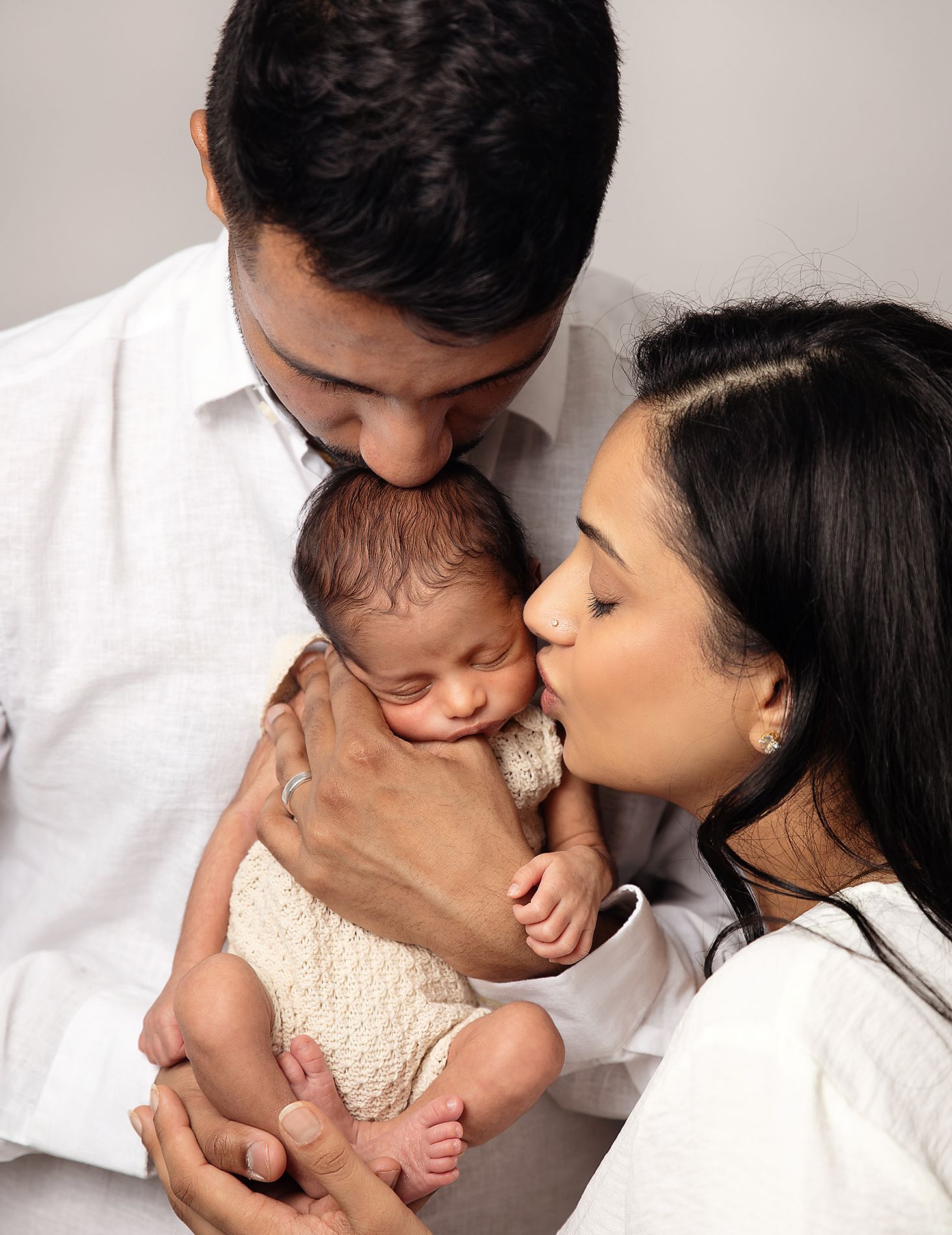 Parents lovingly kiss their newborn baby wearing a cream-colored outfit in a tender family portrait. During a newborn photoshoot in Sidcup