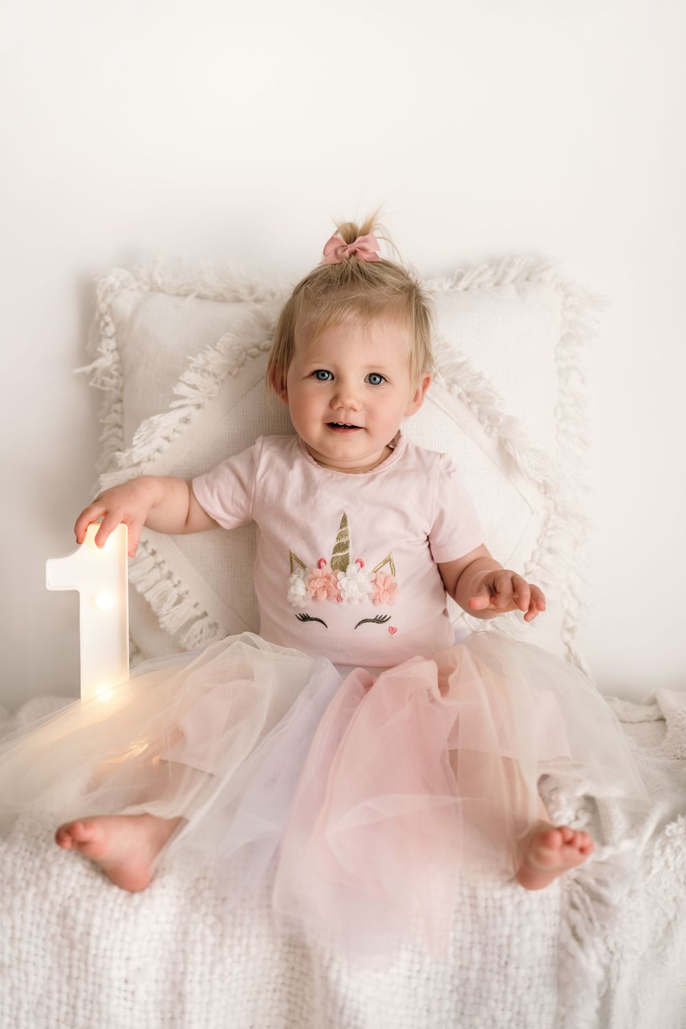 Baby in pink tulle dress with unicorn shirt sits on white bedding next to illuminated number one birthday decoration.