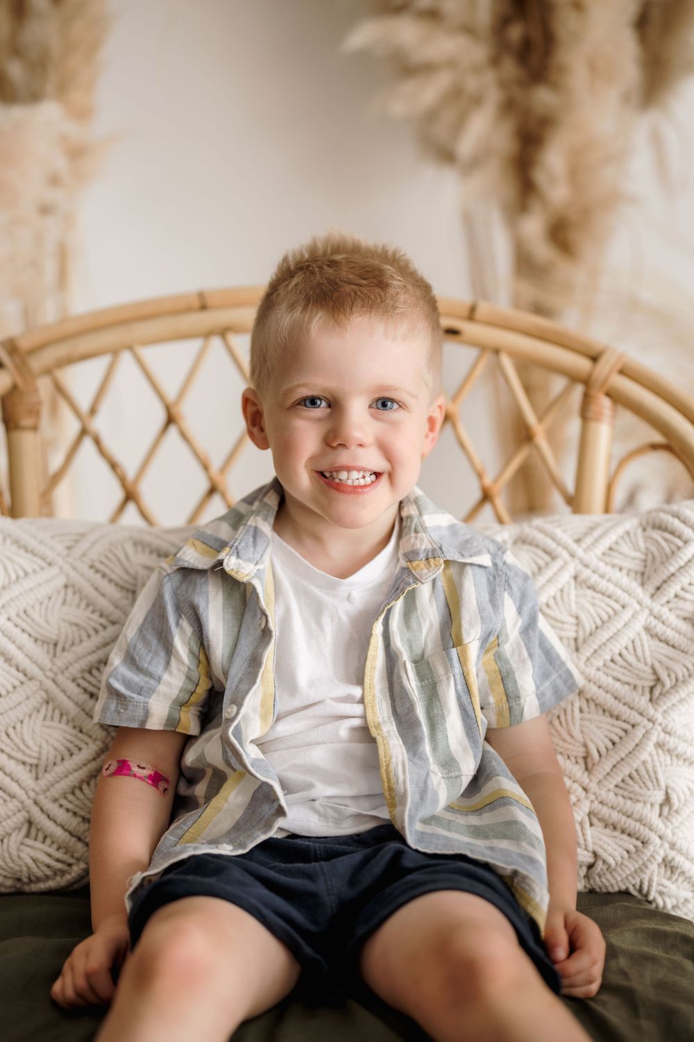 Child sits on textured cushions wearing striped button-down shirt over white tee and navy shorts in warm indoor setting.