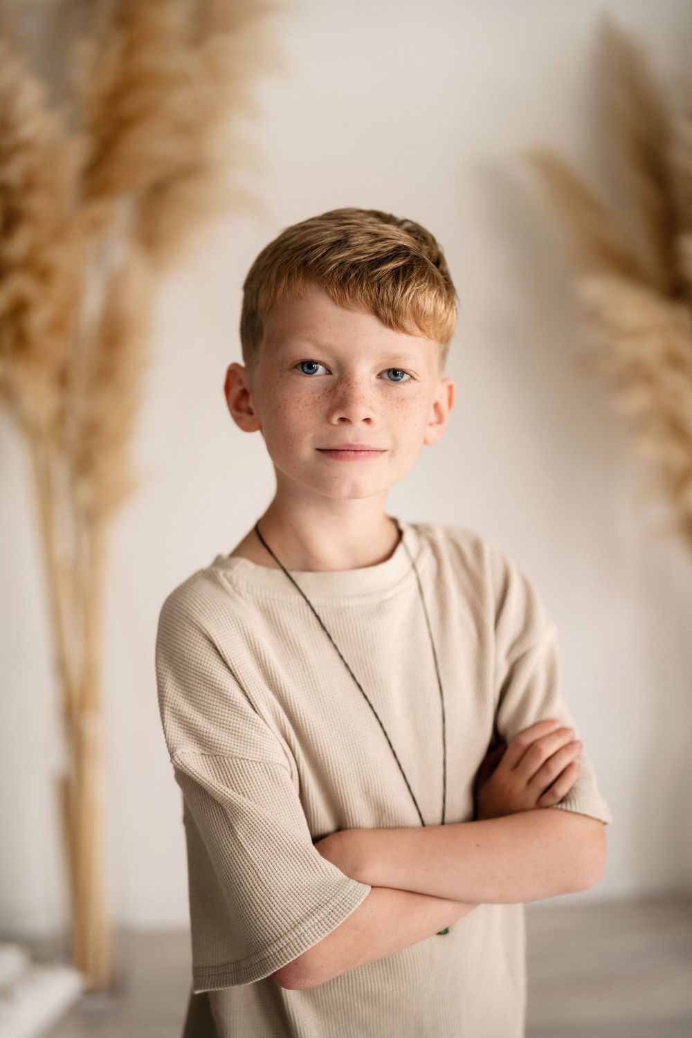 Portrait of child in beige sweater standing with arms crossed against light background with pampas grass decor.