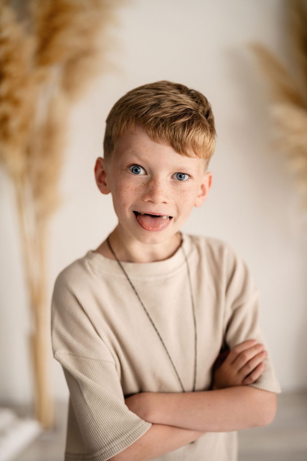 A child wearing a beige sweater stands with arms crossed and an excited expression against a light background with pampas grass.