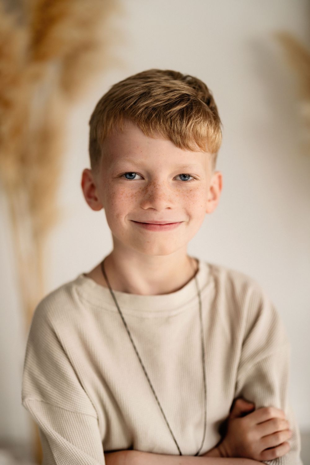 Portrait of a smiling child in a beige sweater with arms crossed against a soft neutral background with pampas grass.