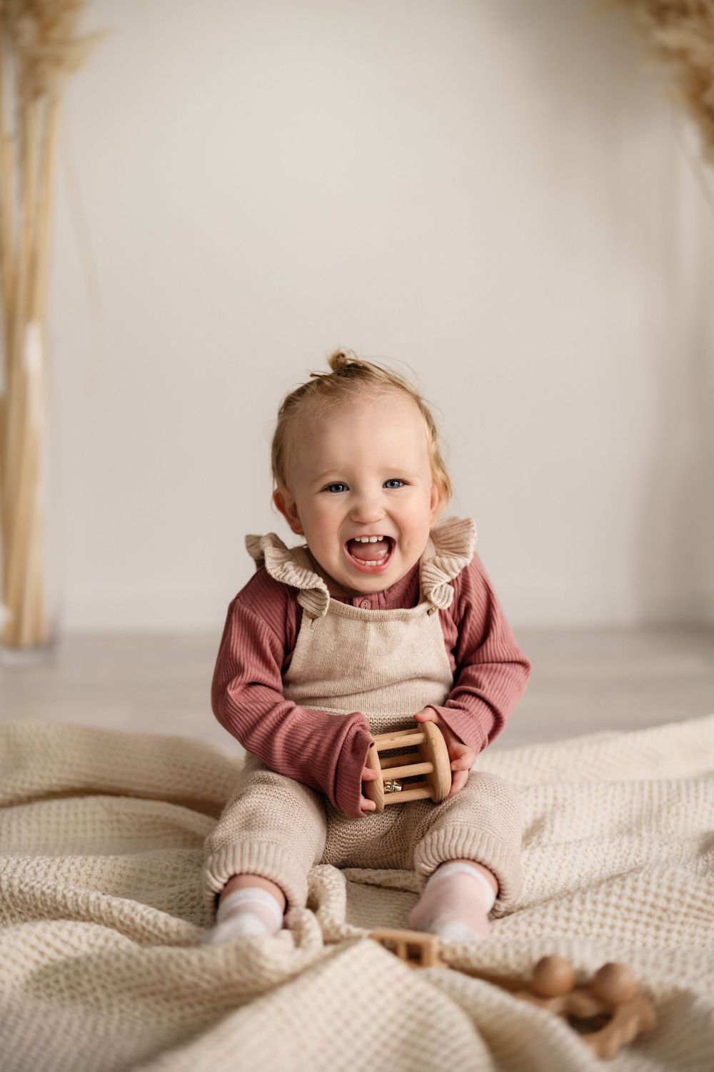 Toddler in rust-colored shirt and beige overalls sits cheerfully on cream knit blanket with wooden toys nearby.