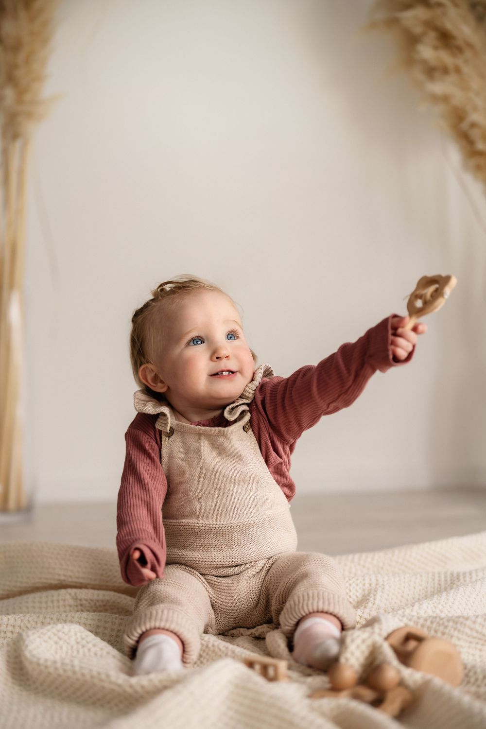 Small child in beige overalls and maroon shirt sits on blanket reaching up with wooden toy in hand.