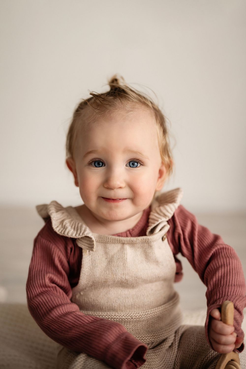 Baby wearing a beige ruffle-collar romper and burgundy sweater smiles while sitting against neutral background.