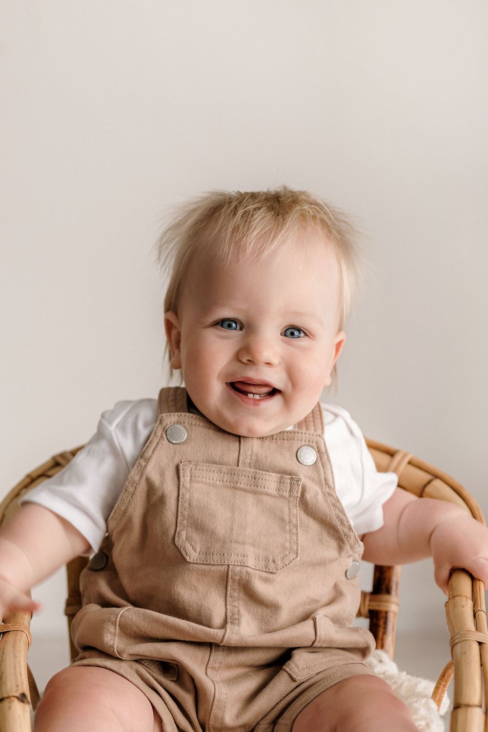 Happy baby wearing tan overalls and white shirt sits in wooden chair against neutral background.