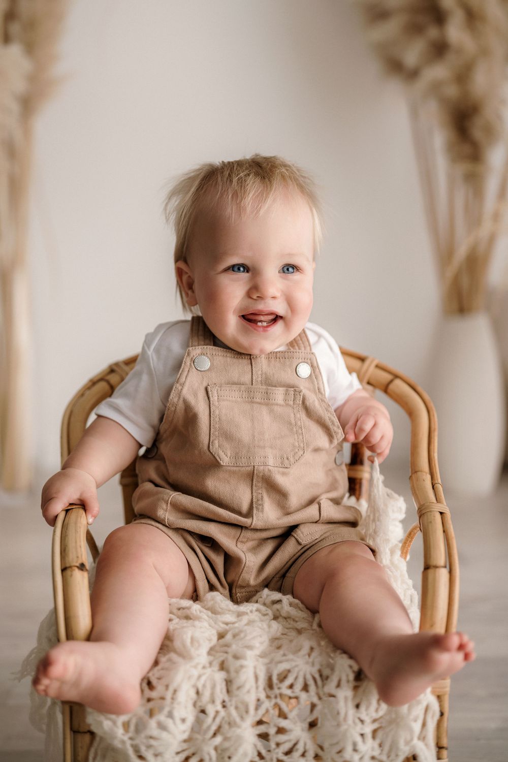 A happy baby wearing tan overalls sits in a wicker chair with a light-colored throw blanket.