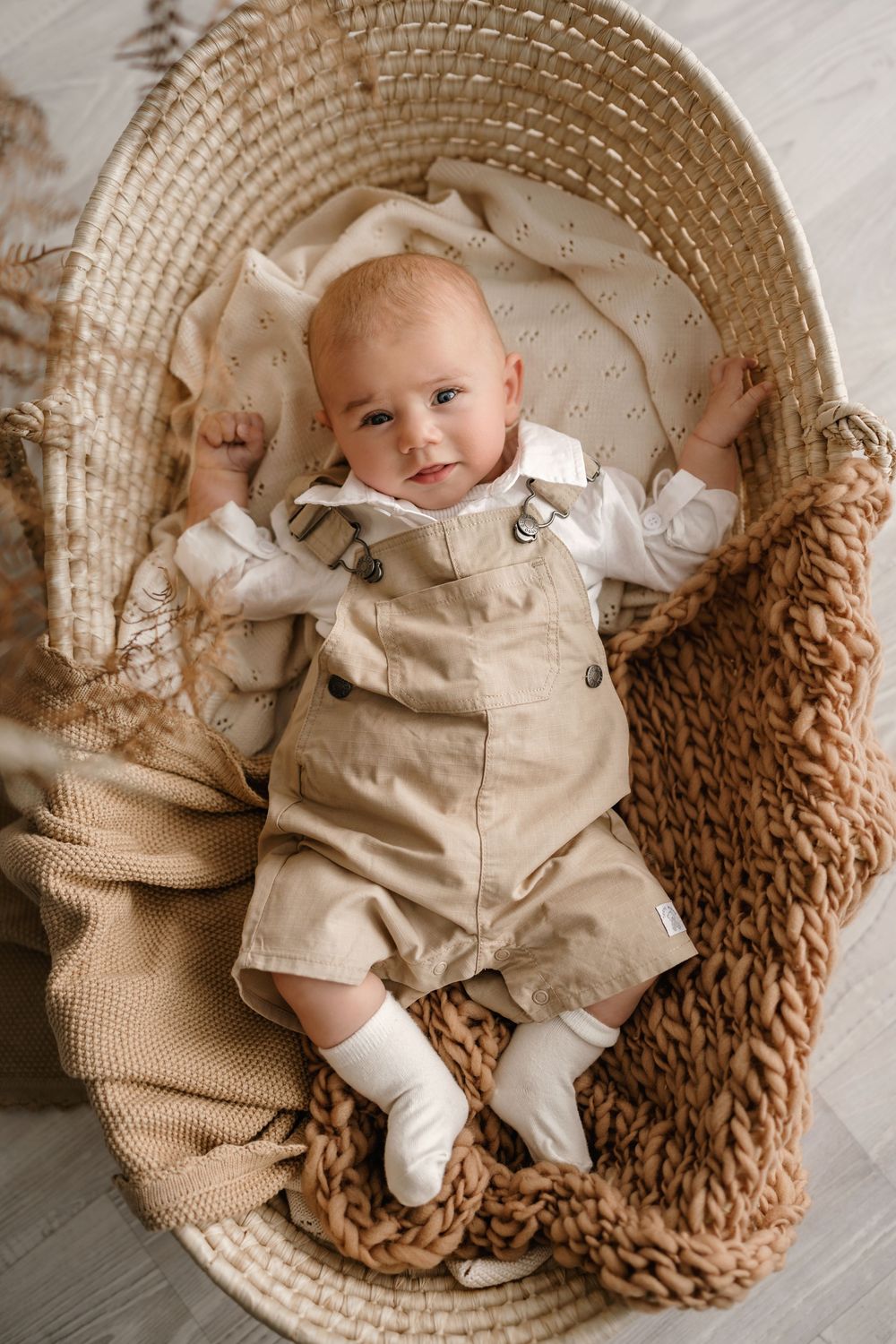 Baby in beige romper outfit lying in woven basket with knit blankets and textured decor in neutral tones.